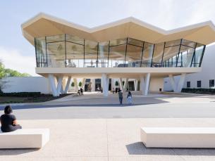 An outdoor setting that shows the façade of a contemporary building. A large walkway with white concrete, block-style benches is in the foreground. The building is raised more than 12 feet off the ground and sits on V-shaped stilts. The front of the building is an all-glass façade and people can be seen inside. The roofline is in a zig-zag shape. The colors are all white and cream with black window panes.