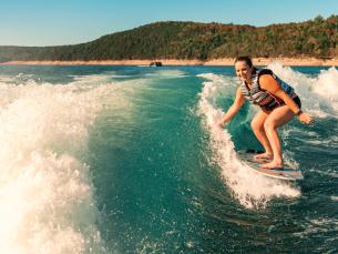 A lake setting near Mountain Home, Arkansas. Blue sky and green-tree hills are seen in the background. Blue water and white wake fill the rest of the photograph. A girl is seen on a board surfing in the wake. Photo source: Magnetic Creative.