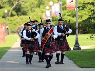 A traditional Scottish bagpiping band is leading a procession of graduates down the sidewalk on the Lyon College campus. All wearing kilts, the man in front is wearing a sash and carrying a baton. Three pipers are walking behind and playing, as well as a drummer. Street lamps are adorned with red and blue banners.