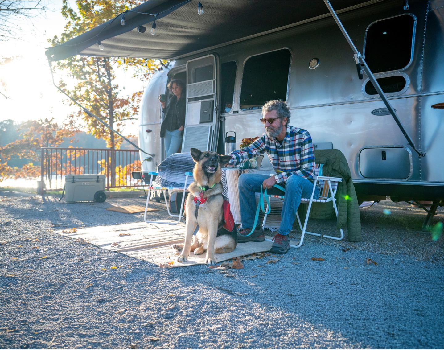 Man sitting by a camper petting a dog, with trees in the background.