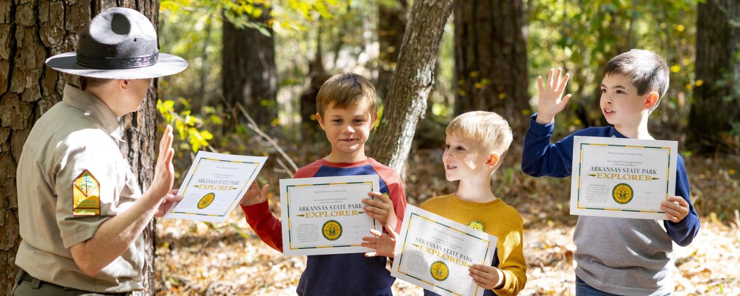 Park ranger congratulates three kids holding certificates in a forest.
