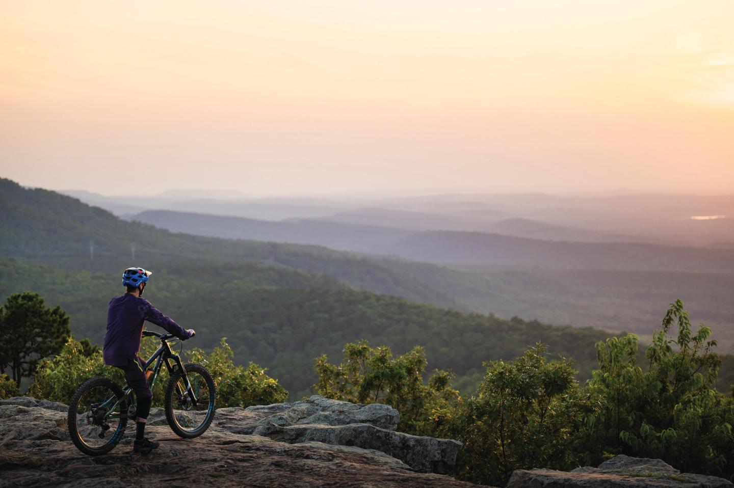 Cyclist on a hilltop at sunset, overlooking a vast, misty landscape.