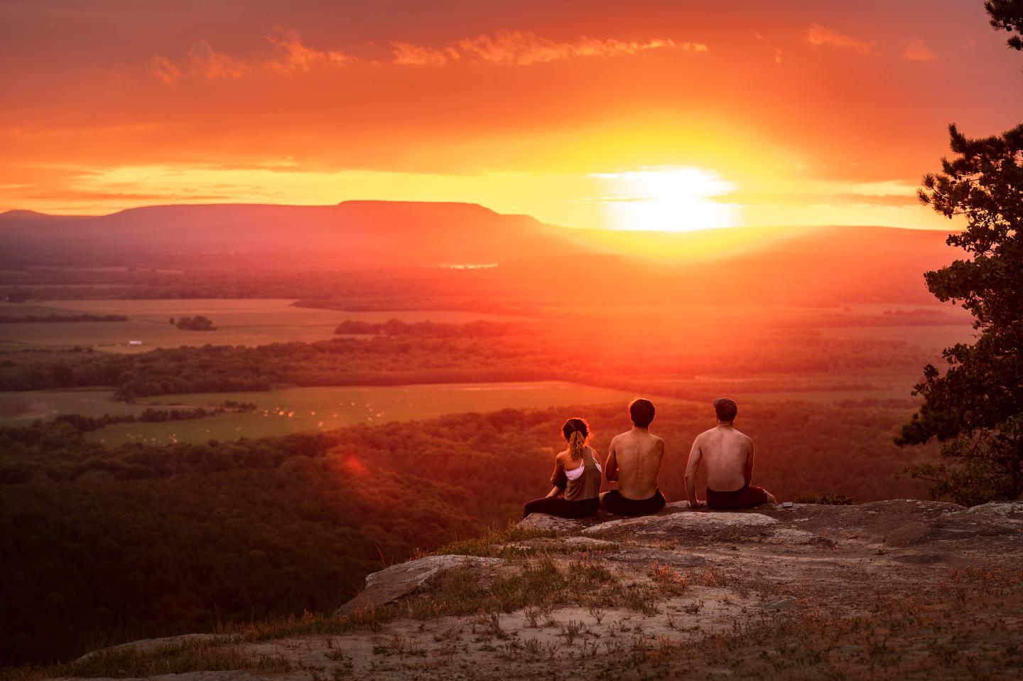 Three people sit on a cliff edge watching a vibrant sunset over a vast landscape.