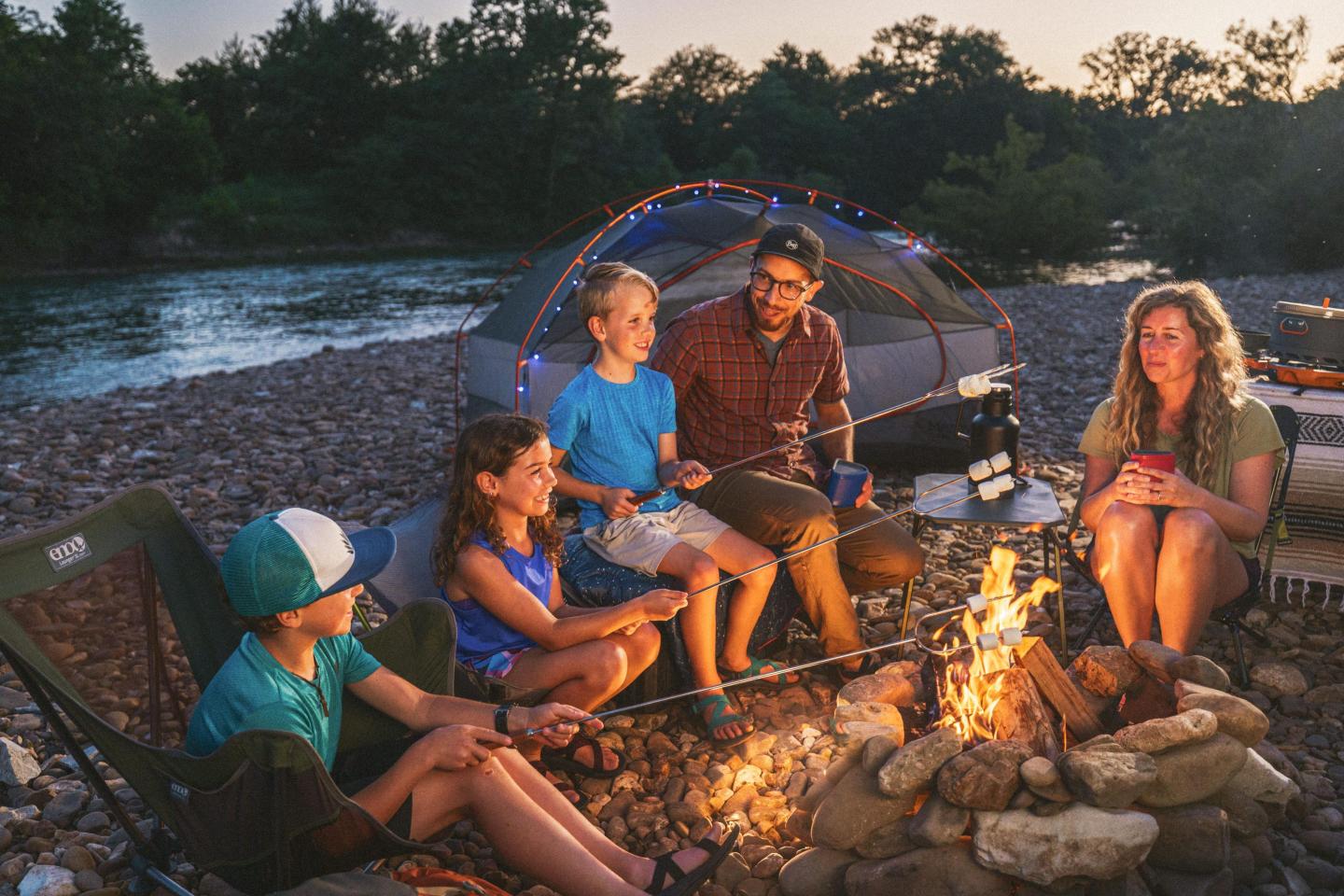 Family gathered around a campfire by a river at dusk, with a tent in the background.