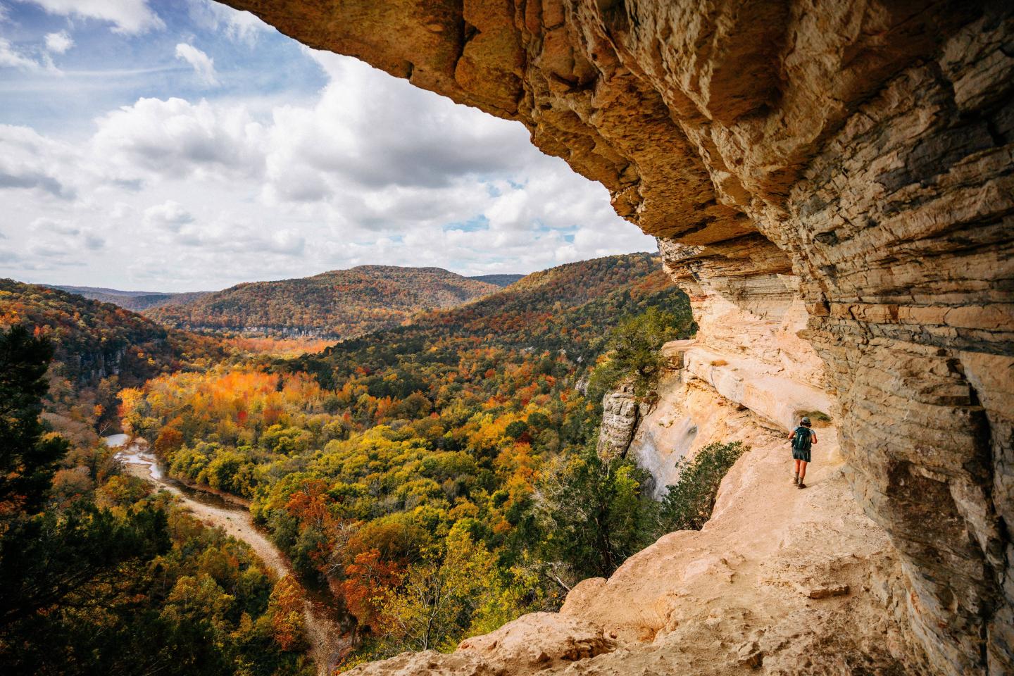 Cliff side view with person hiking, overlooking colorful autumn forest and river.