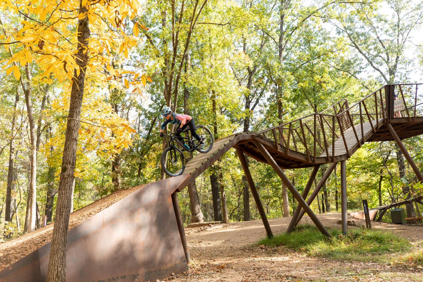 Cyclist on wooden ramp in forest, trees with autumn leaves.