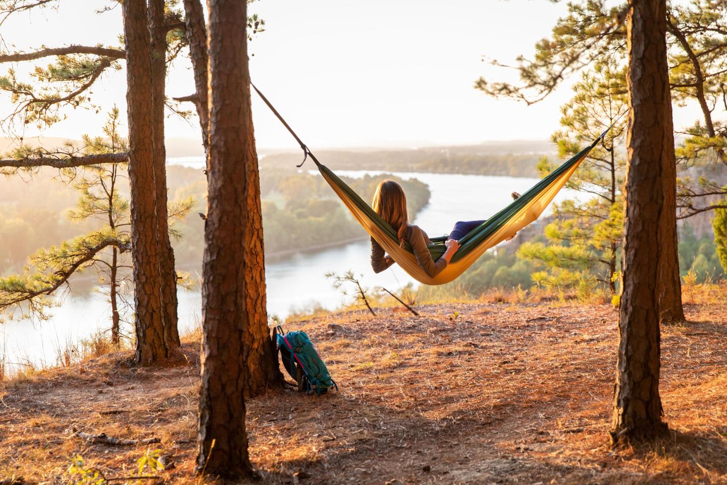 Person relaxing in a hammock between trees overlooking a river at sunset.