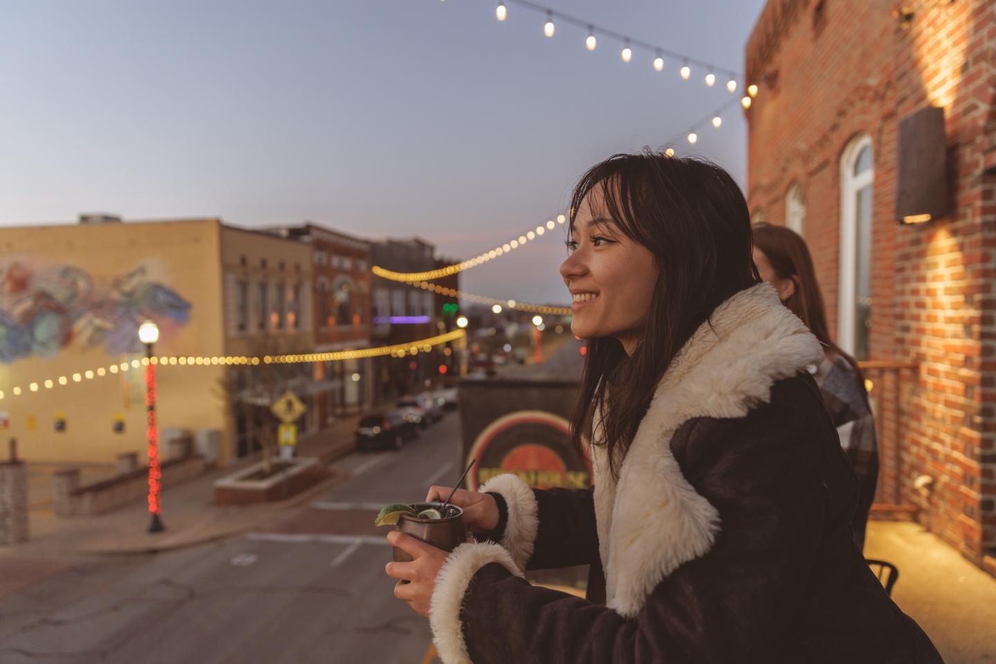 Woman smiling on a balcony at sunset, holding a camera, with city street lights in view.