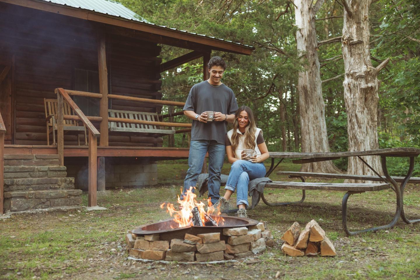 Couple by a campfire outside a rustic cabin, surrounded by trees.
