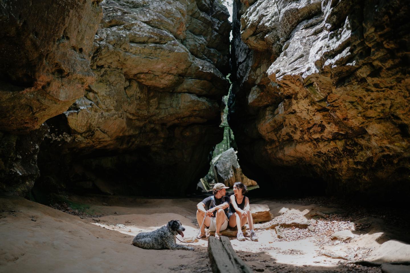 Two people and a dog sit between large rock formations, sunlight casting shadows.