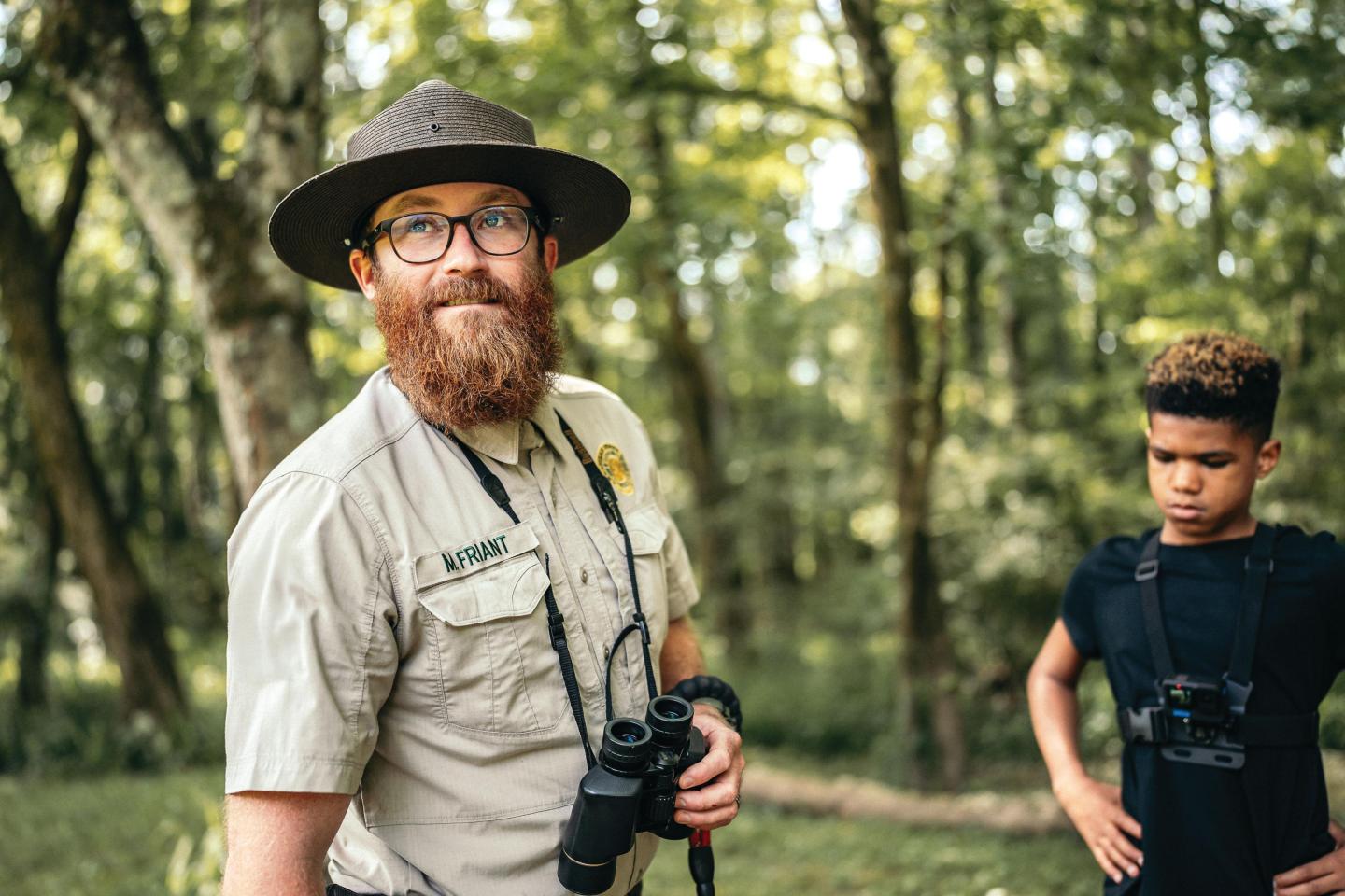 Park ranger with binoculars and boy in wooded area.