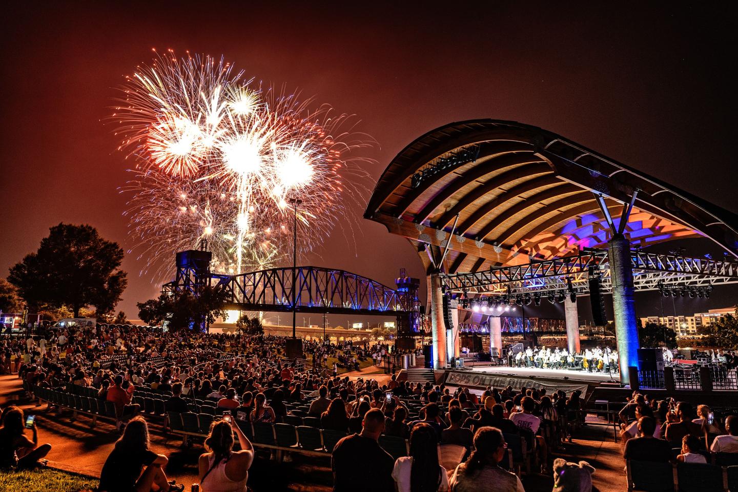 Fireworks over a concert at night, with a crowd under a lit amphitheater.