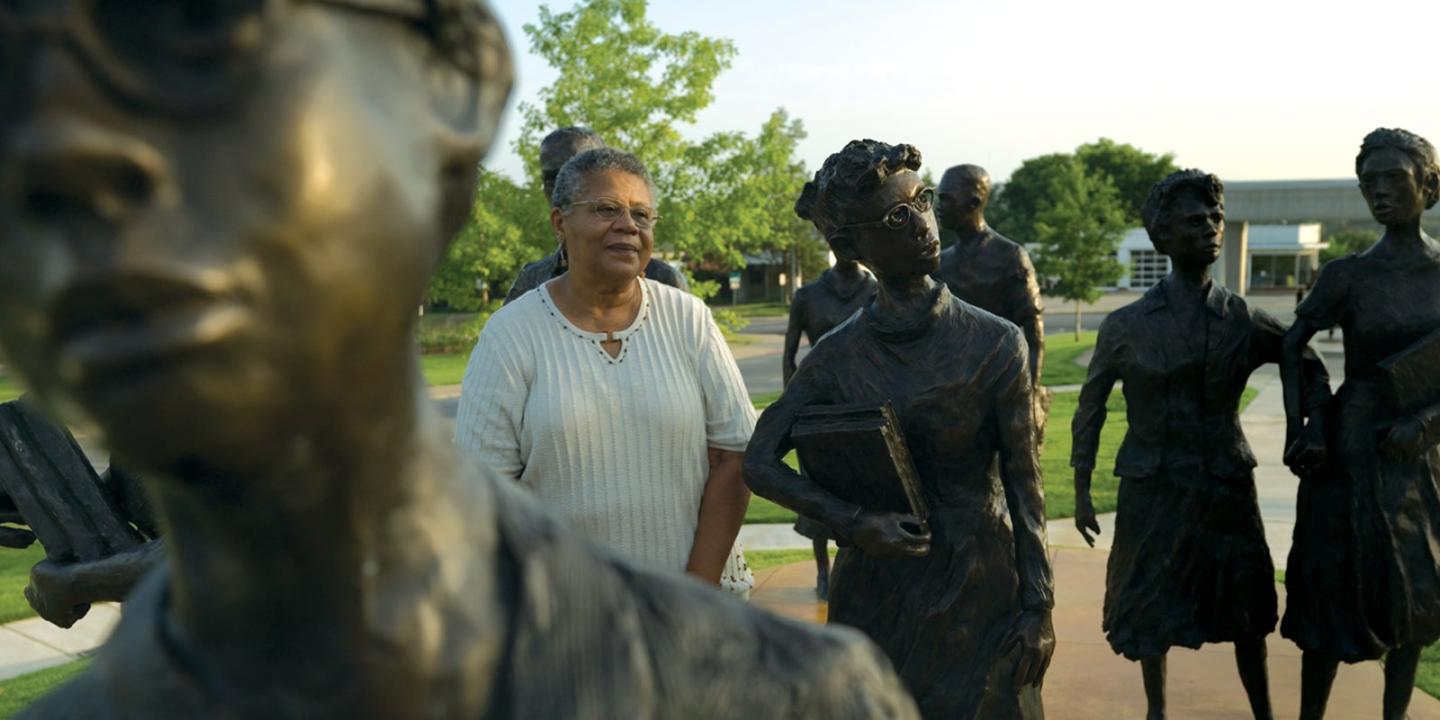 Smiling woman stands among bronze statues in a park setting.