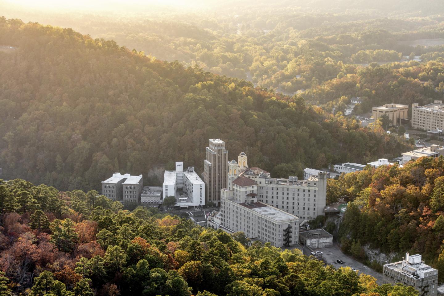 Aerial view of buildings surrounded by forested hills in soft sunlight.