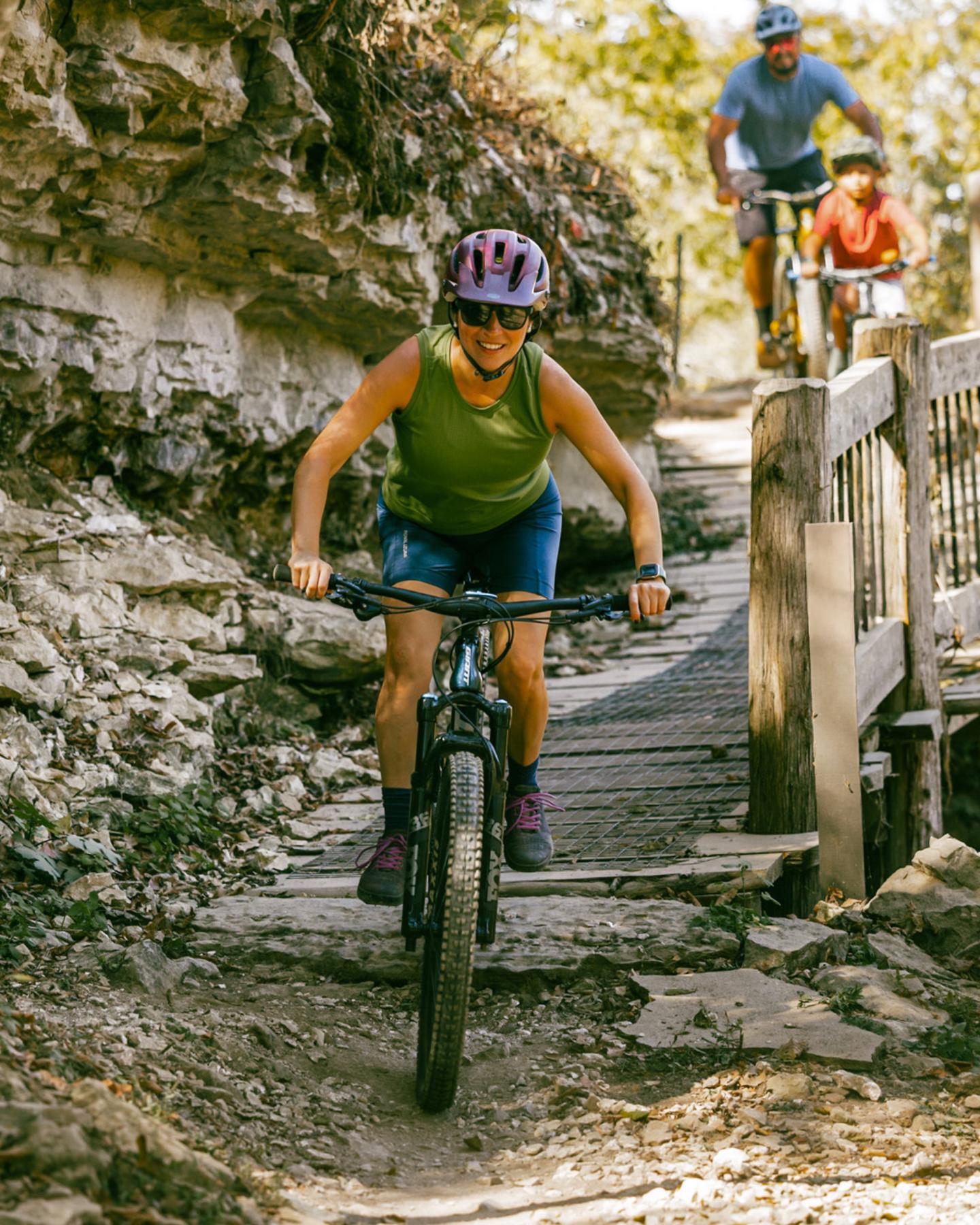 Cyclists ride along a rocky, wooded trail on mountain bikes.