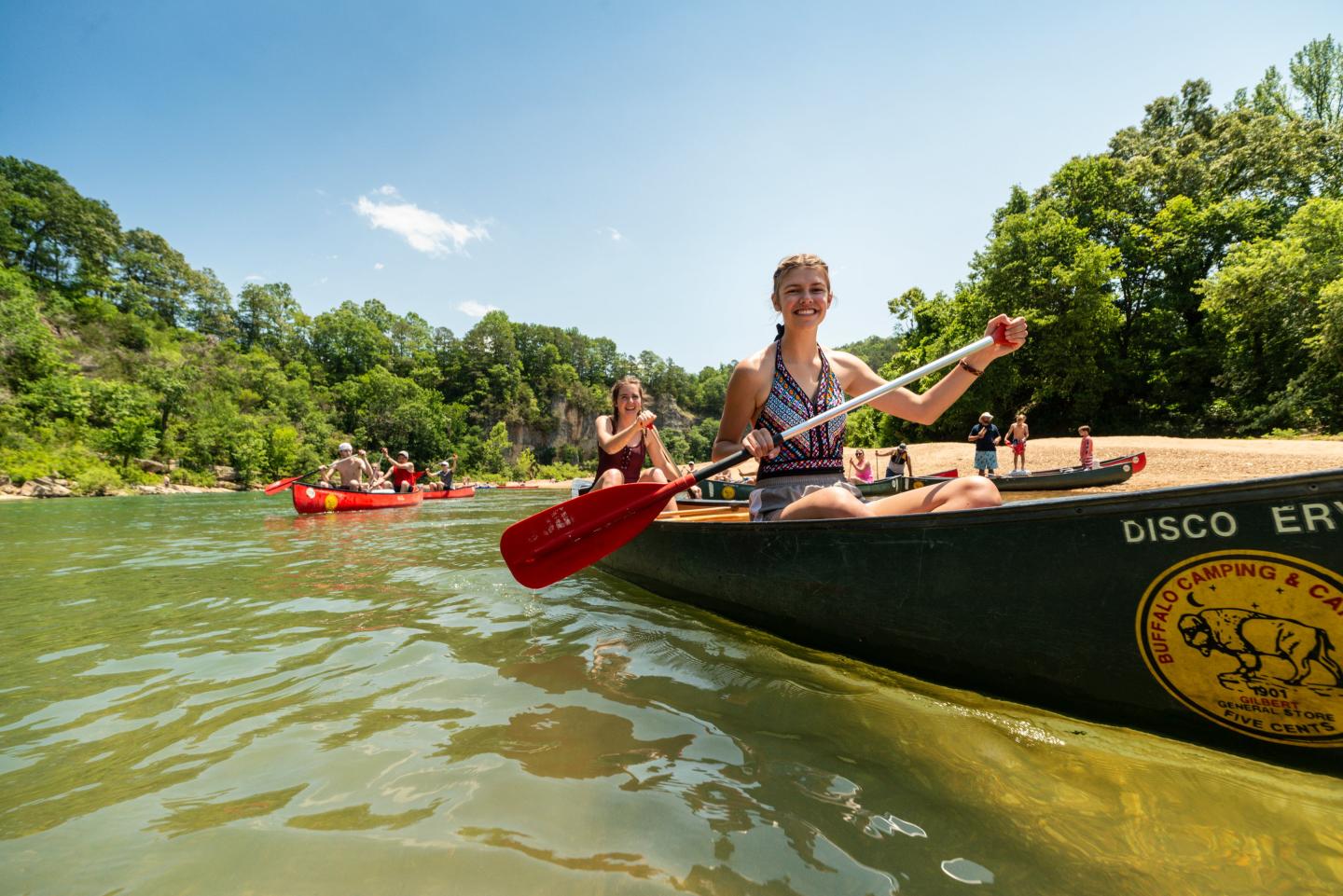 People kayaking on a sunny day, surrounded by green trees on a river.