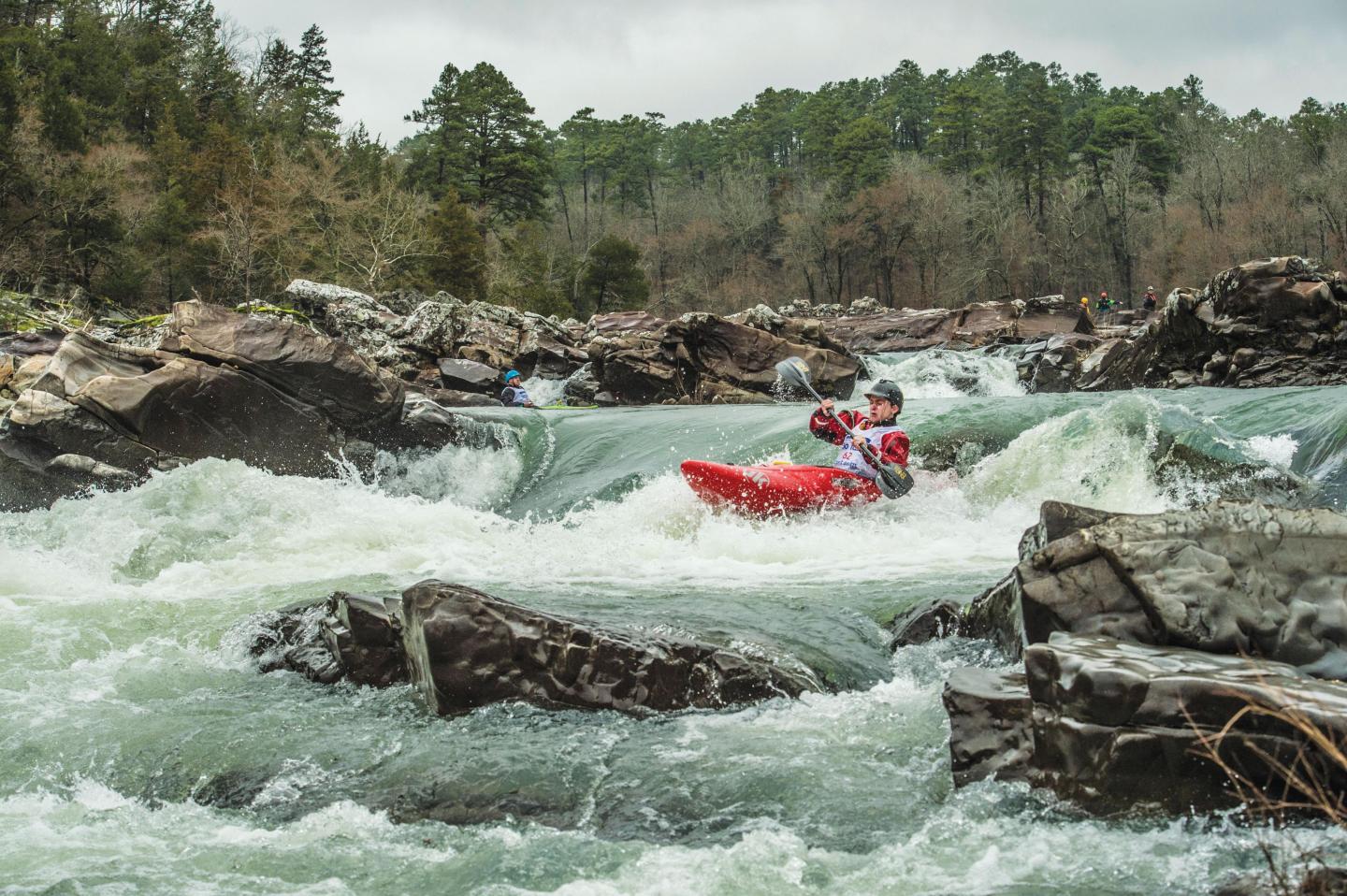 Kayaker in red navigating rapids surrounded by rocks and trees.