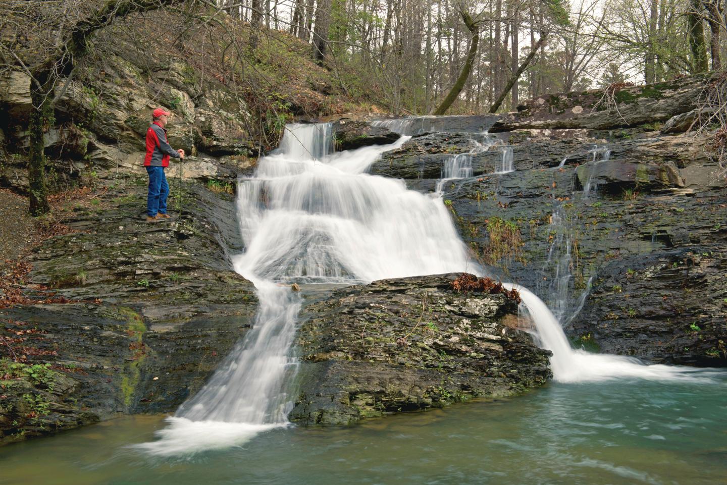Person standing beside a cascading waterfall, surrounded by rocks and trees.