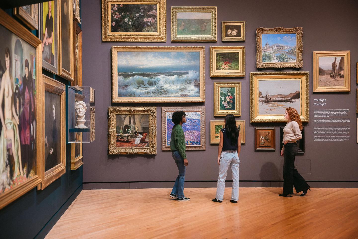 Three people in an art gallery observing paintings on a wall.
