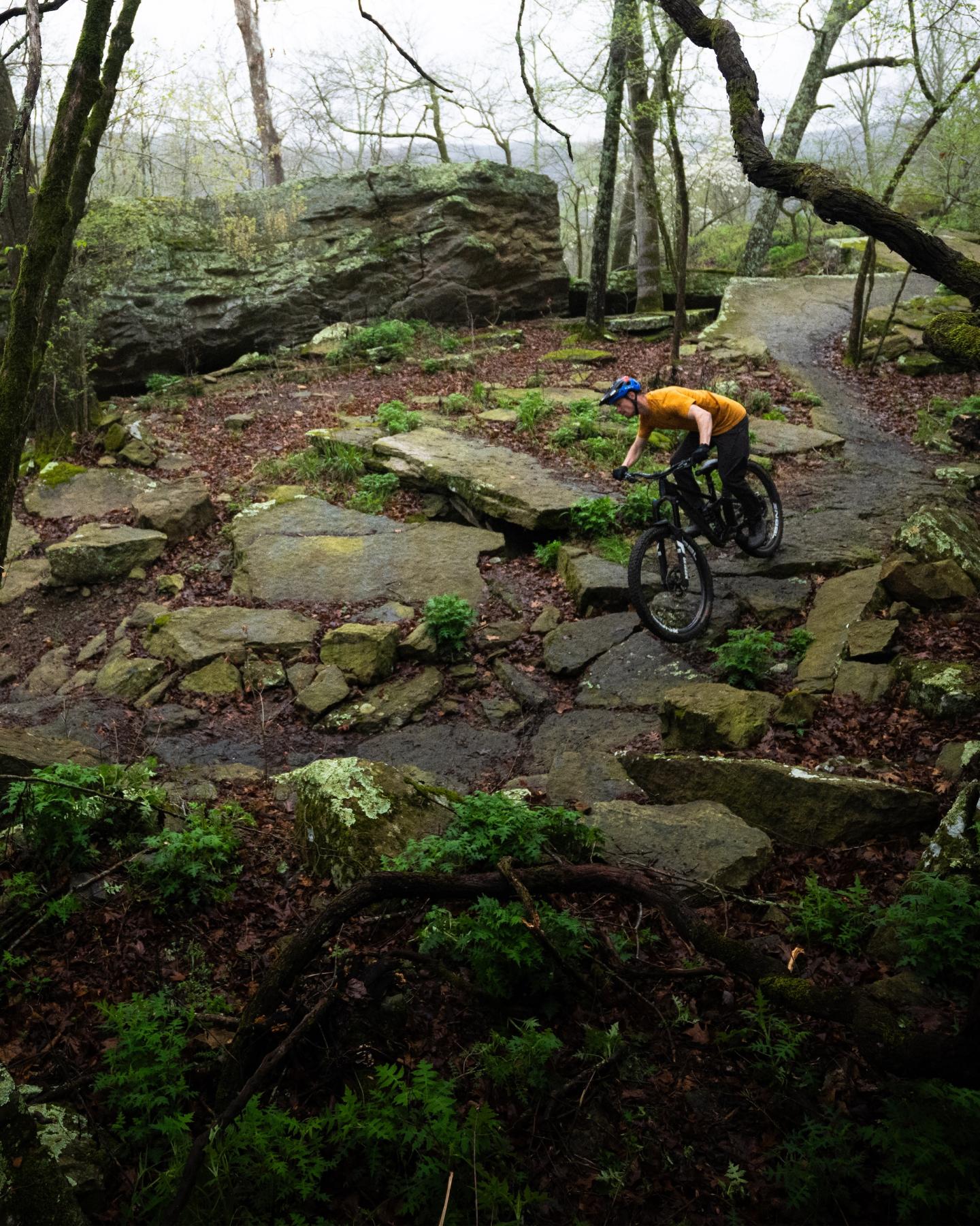 Cyclist on a rocky forest trail, wearing an orange jacket.