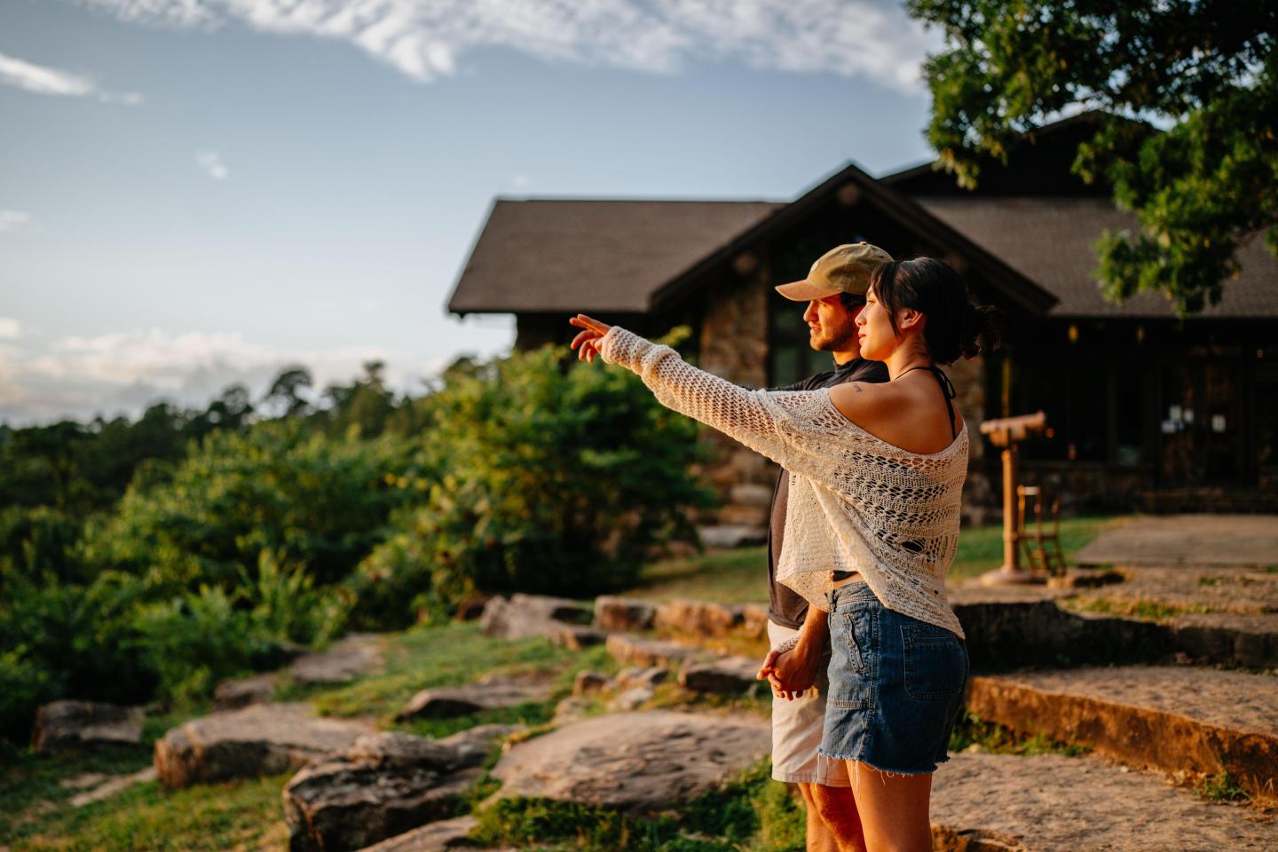 A couple outdoors, one pointing, near a rustic cabin, surrounded by greenery.