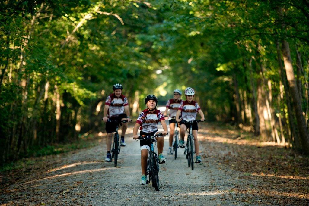 Cyclists ride on a tree-lined dirt path, wearing matching jerseys.