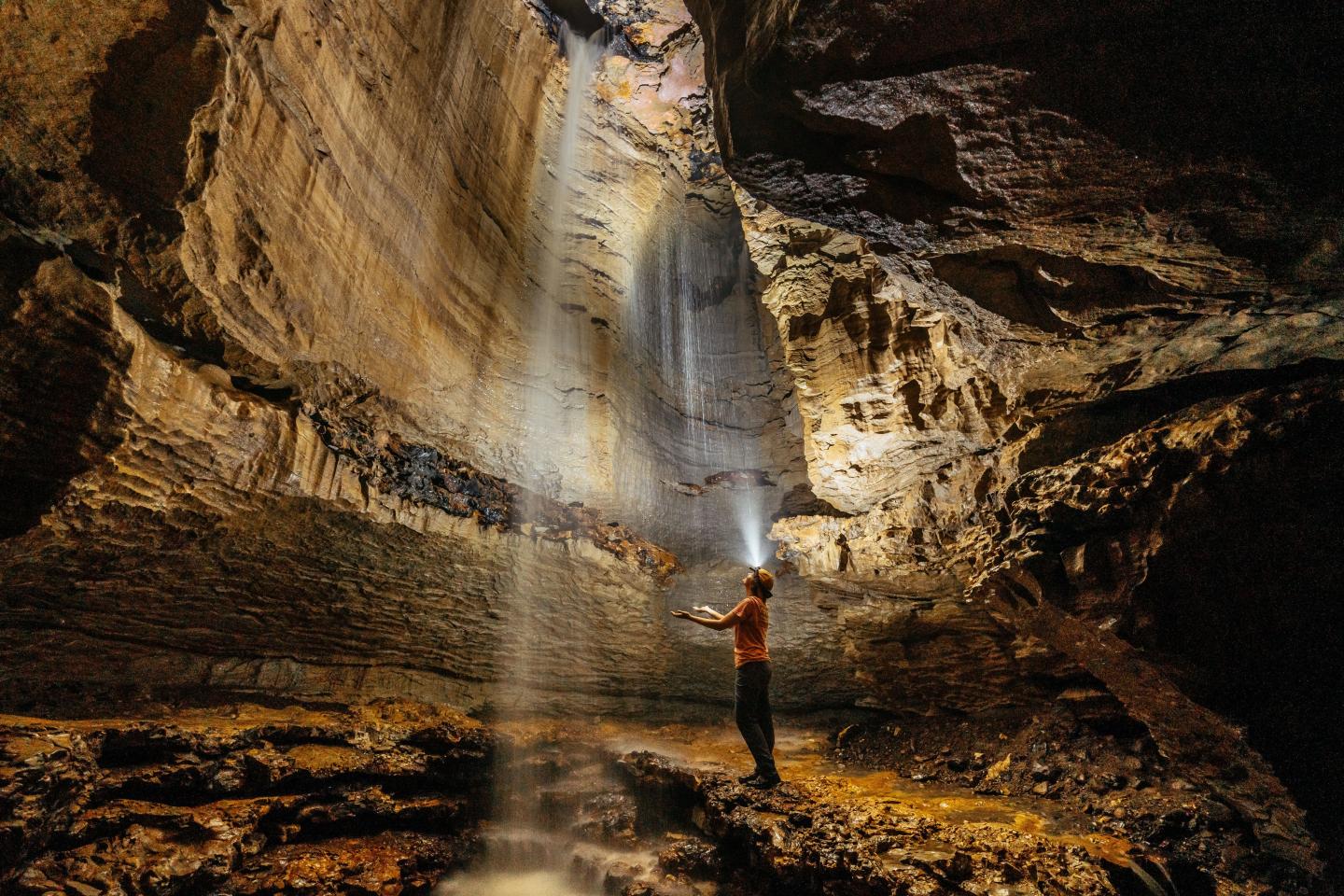 Person standing under a cascading waterfall inside a large, illuminated cave.