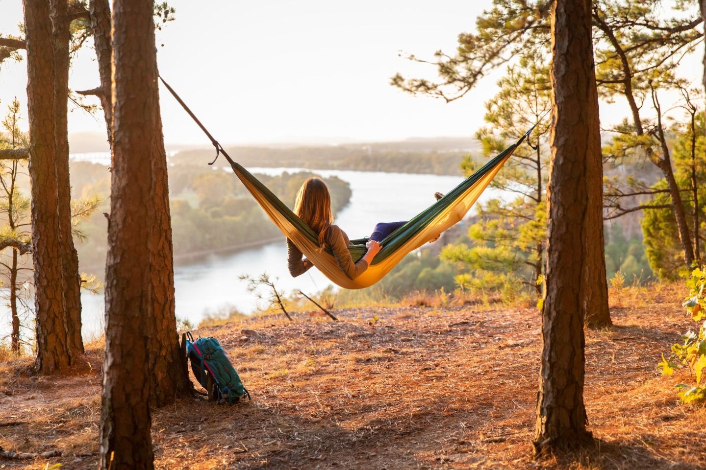 Person relaxing in a hammock between trees overlooking a river at sunset.