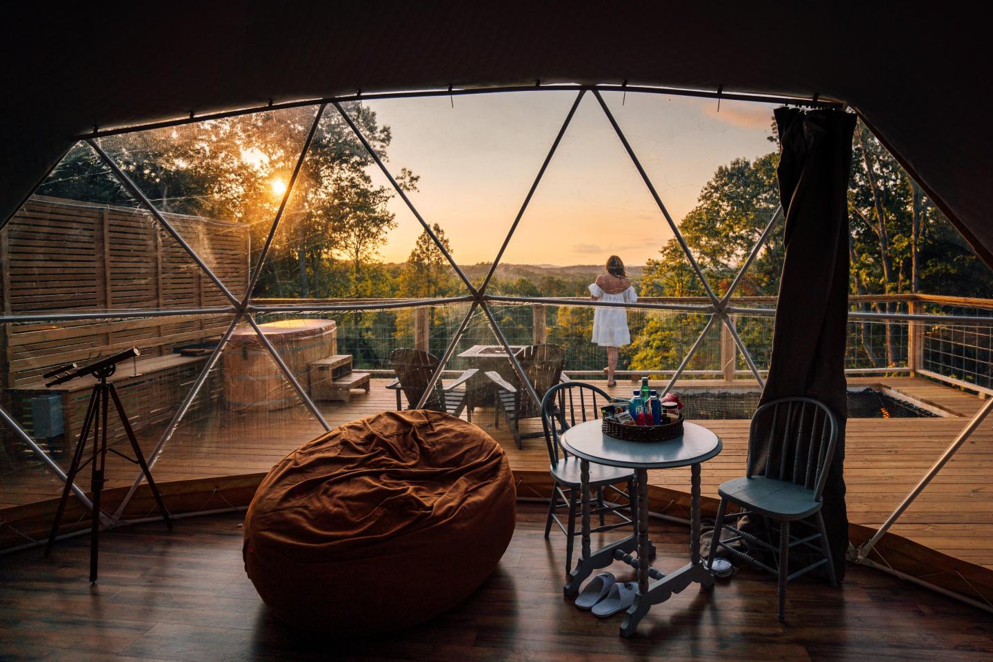 Geodesic dome room with large window, woman on balcony, sunset view.