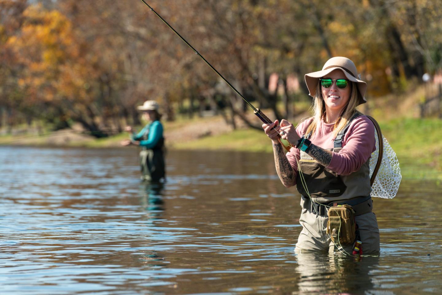 Two people fly fishing in a river on a sunny autumn day, wearing hats and waders.