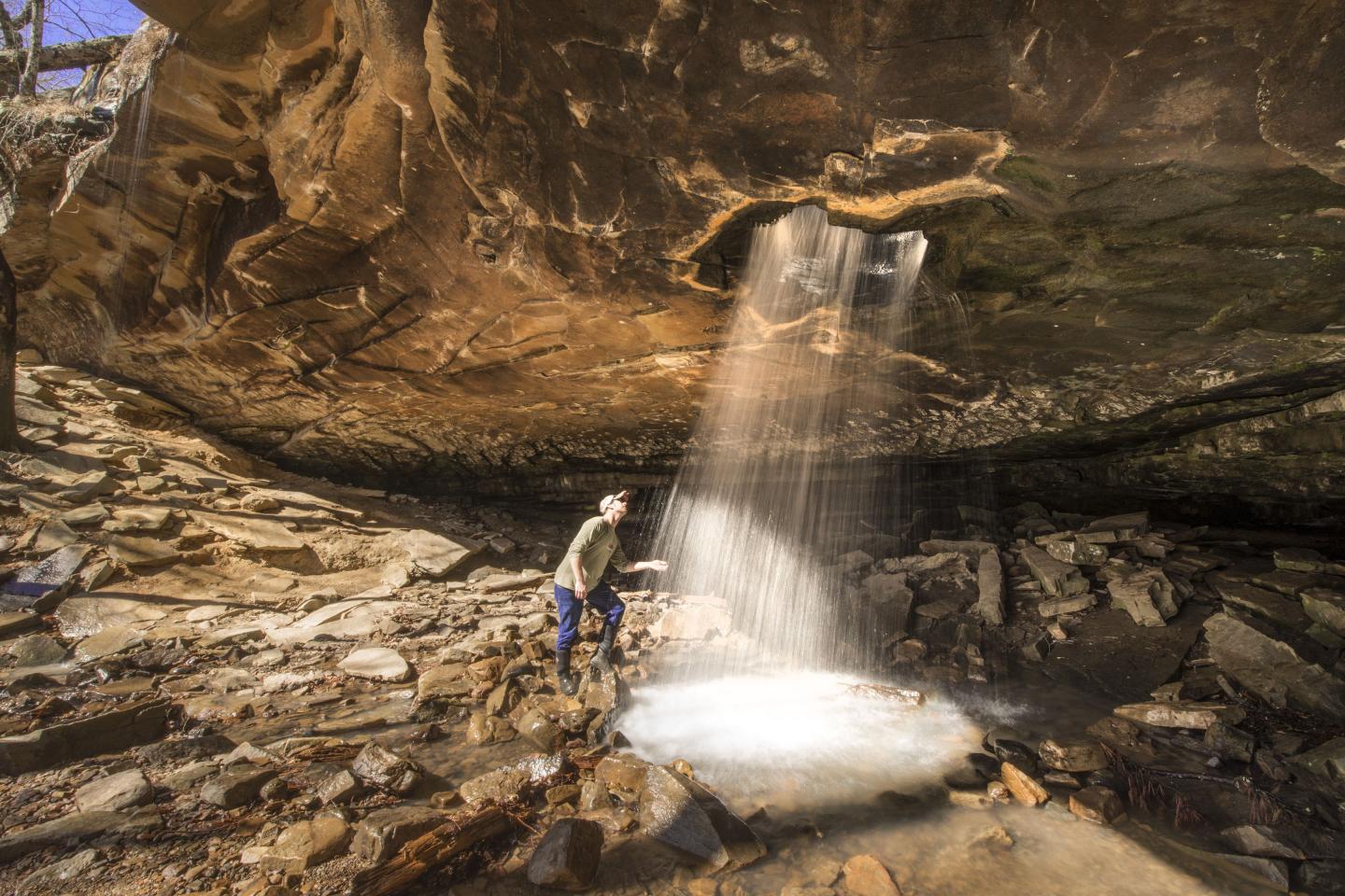 Person standing under cave waterfall, surrounded by rocks and sunlight.