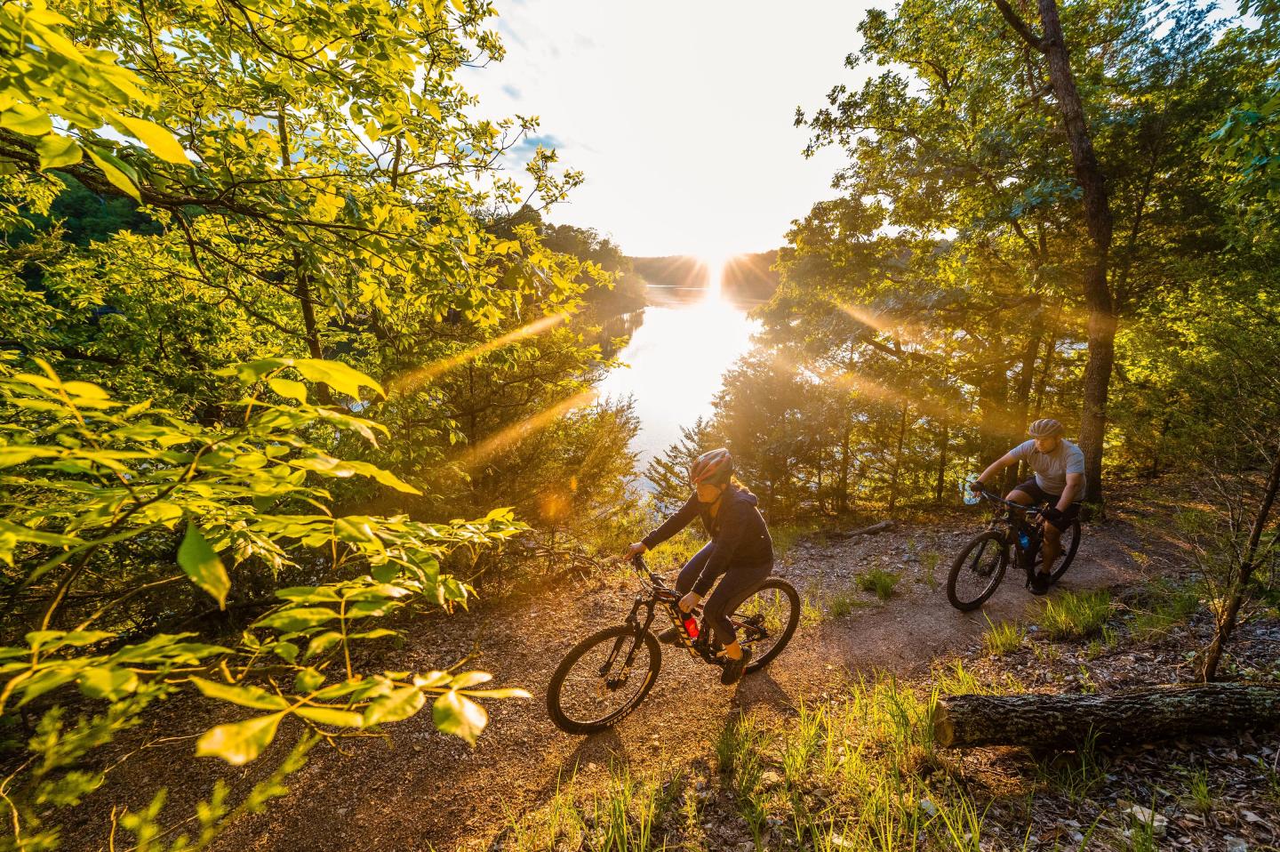 Two cyclists ride on a forest trail by a sunlit lake.