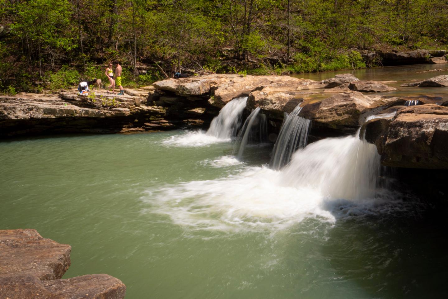 Waterfall flowing into a clear green pool surrounded by rocks and greenery.