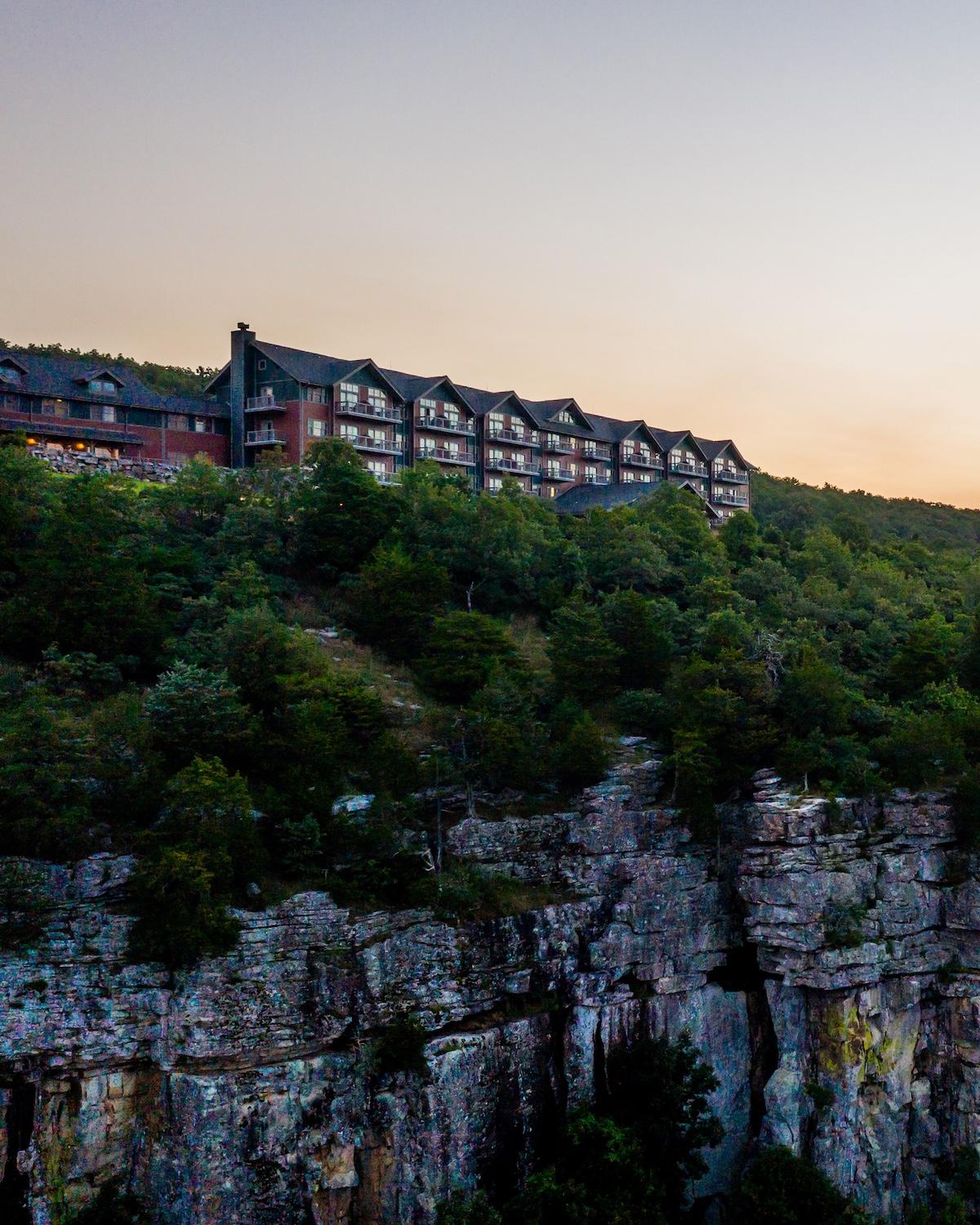 Hotel perched on a cliff, surrounded by lush green trees, under a soft evening sky.