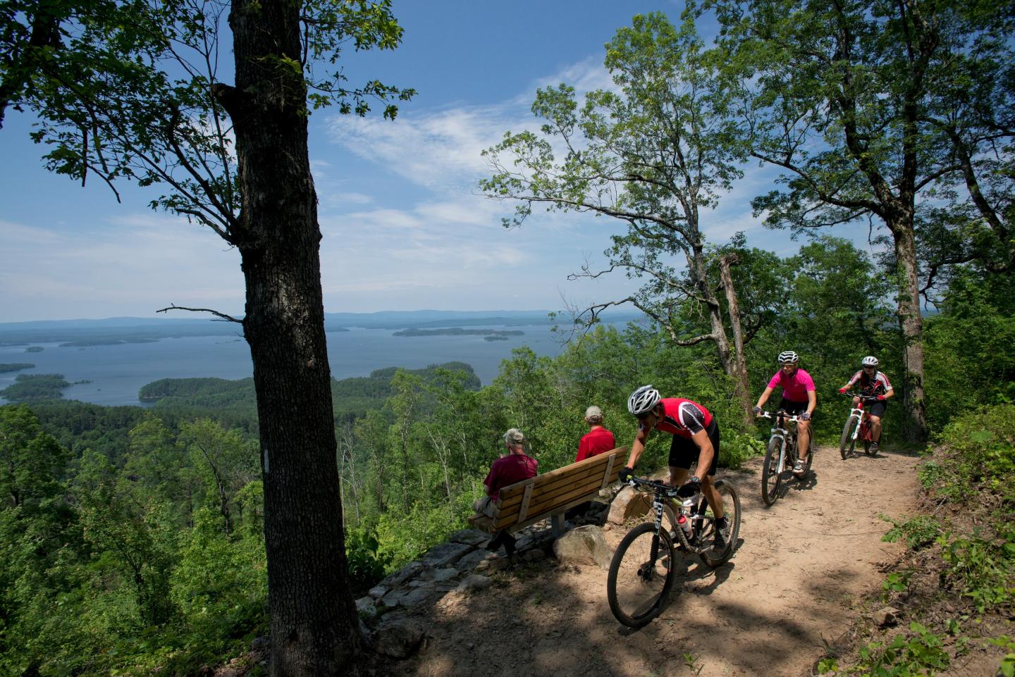 Cyclists ride a forest trail near a bench overlooking a scenic lake view.