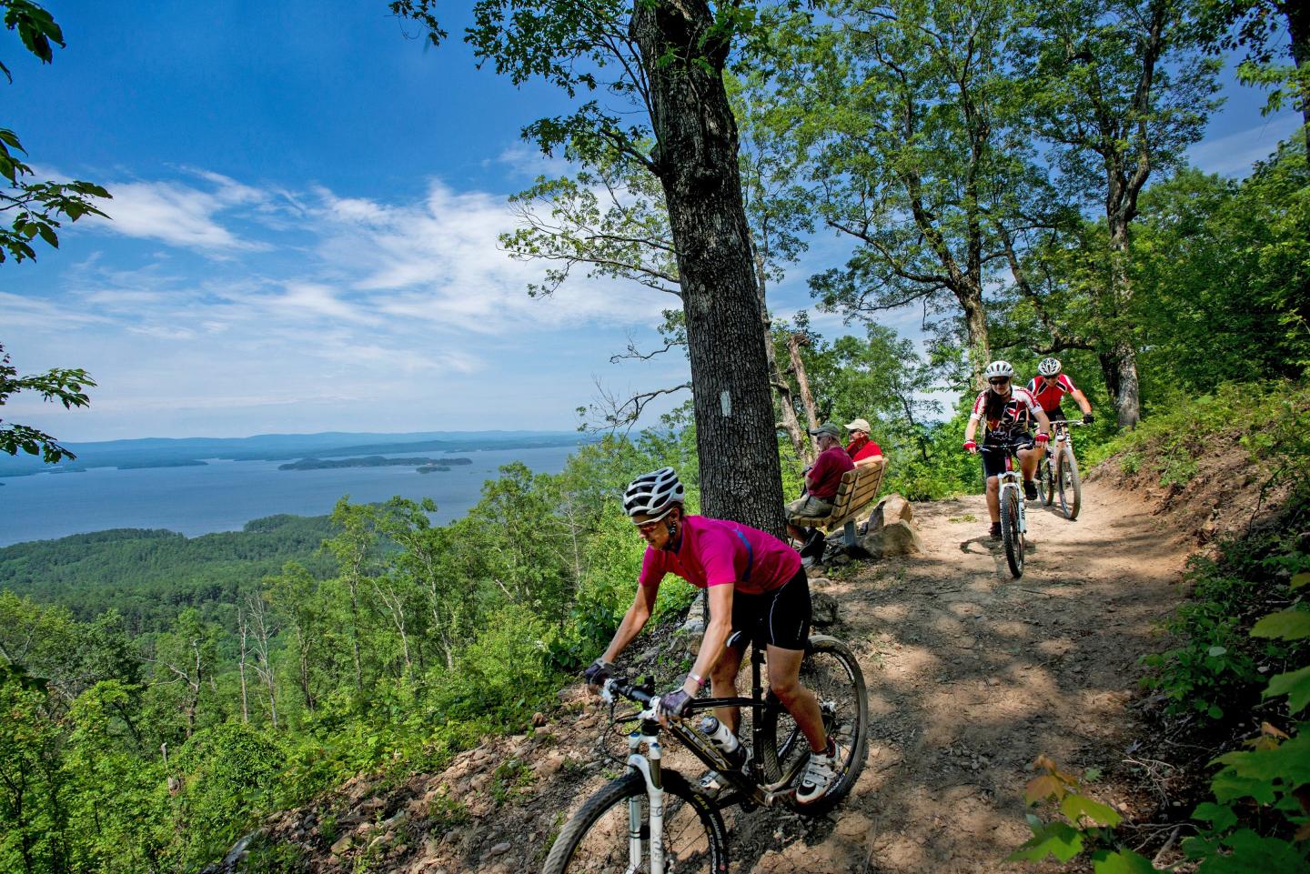 Mountain bikers ride a forest trail overlooking a blue lake on a sunny day.