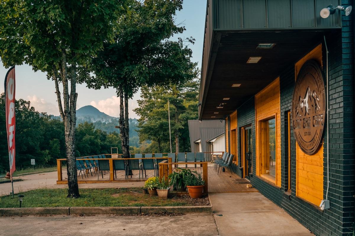 Modern cafe exterior with patio seating and trees, mountains in the background.