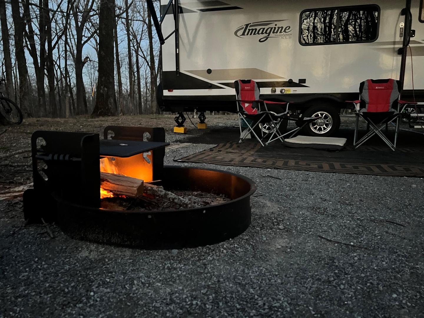 Campfire near RV and chairs in woodland setting at dusk.
