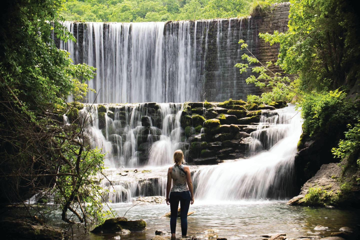 Person standing in front of a cascading waterfall surrounded by lush greenery.