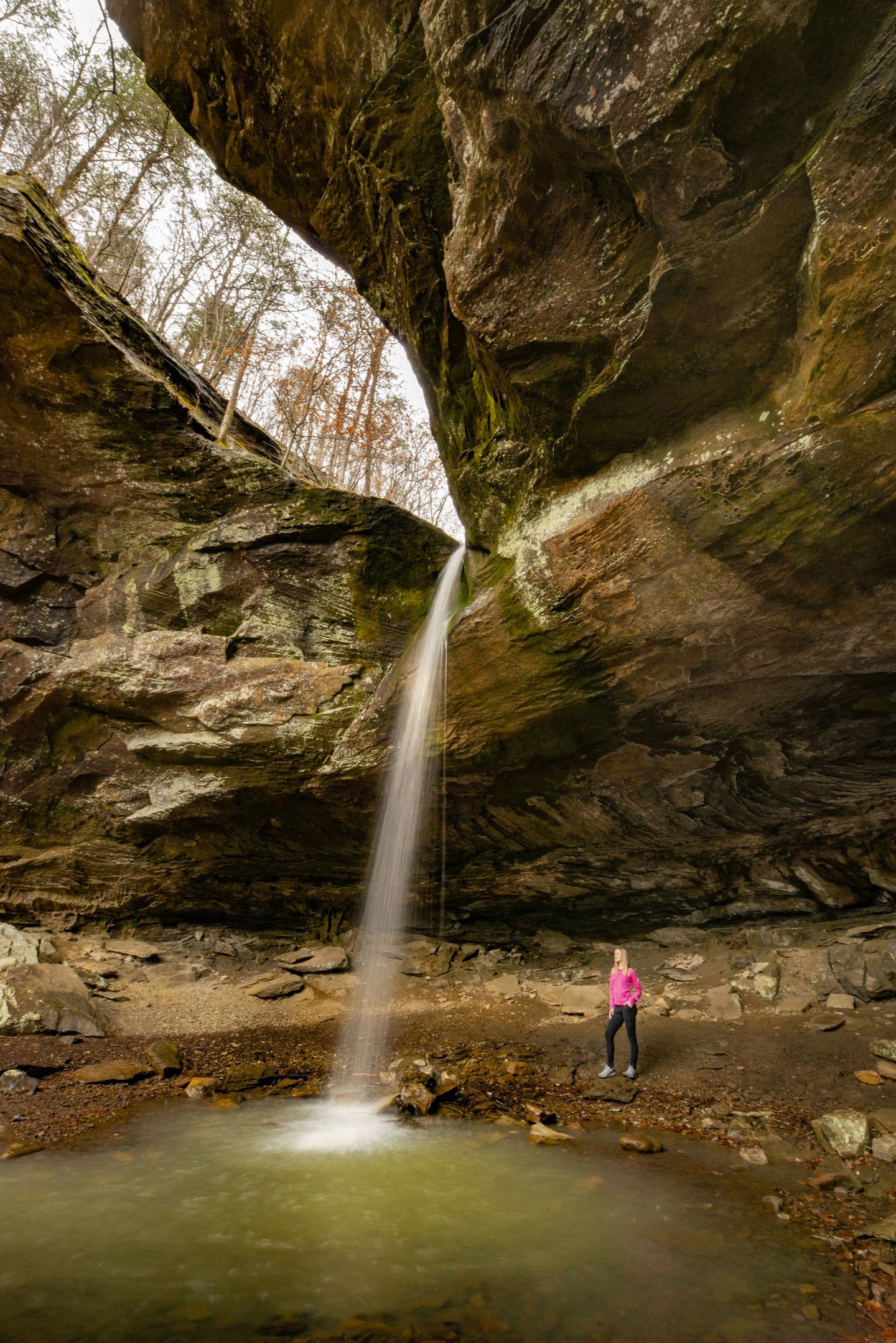 Waterfall in rocky cavern, person in pink jacket stands below.