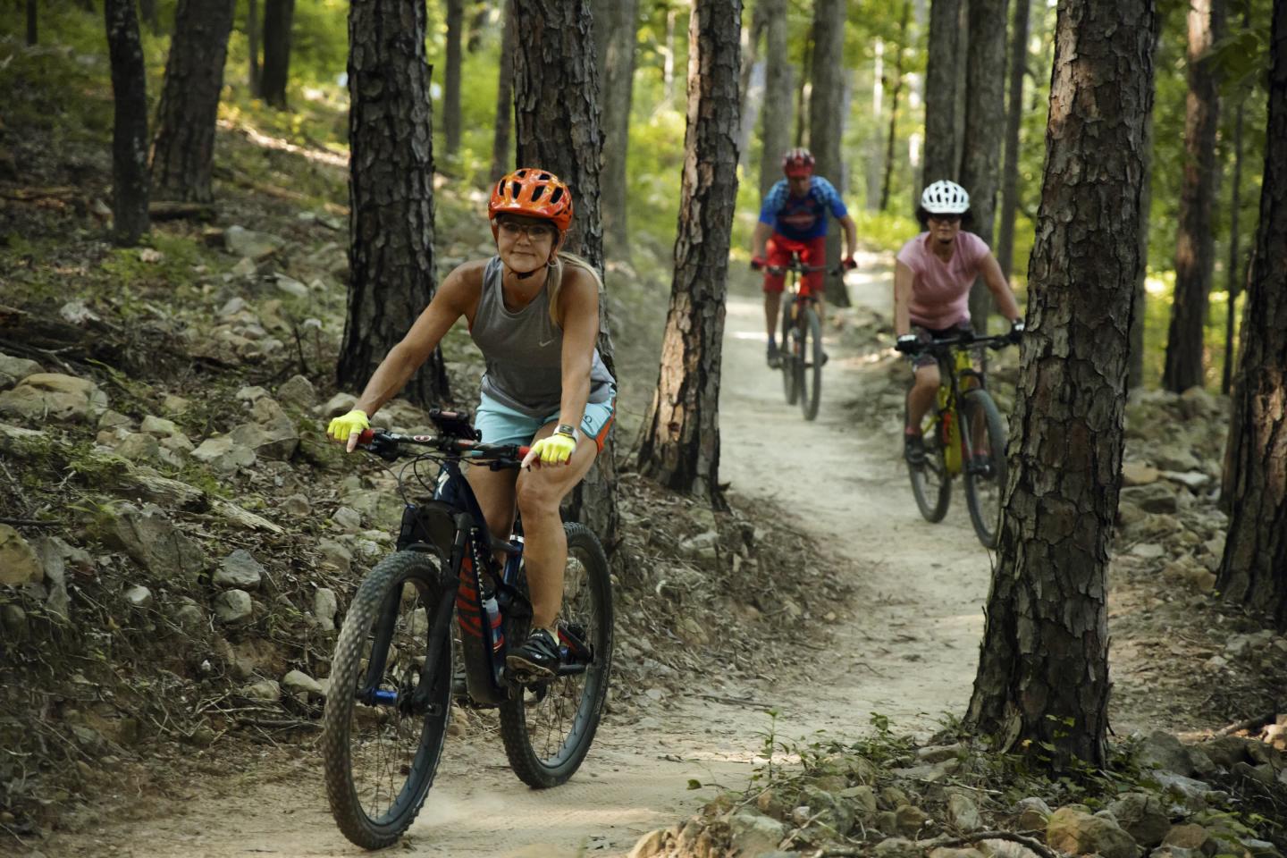 Cyclists on a forest trail, wearing helmets and casual attire.
