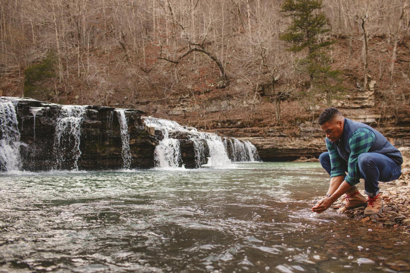 Man washing hands in a river near a small waterfall in a wooded area.