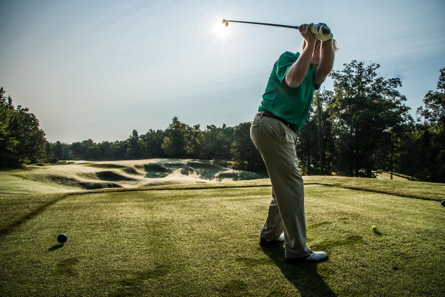 Golfer swings on a sunny course with a pond and trees.
