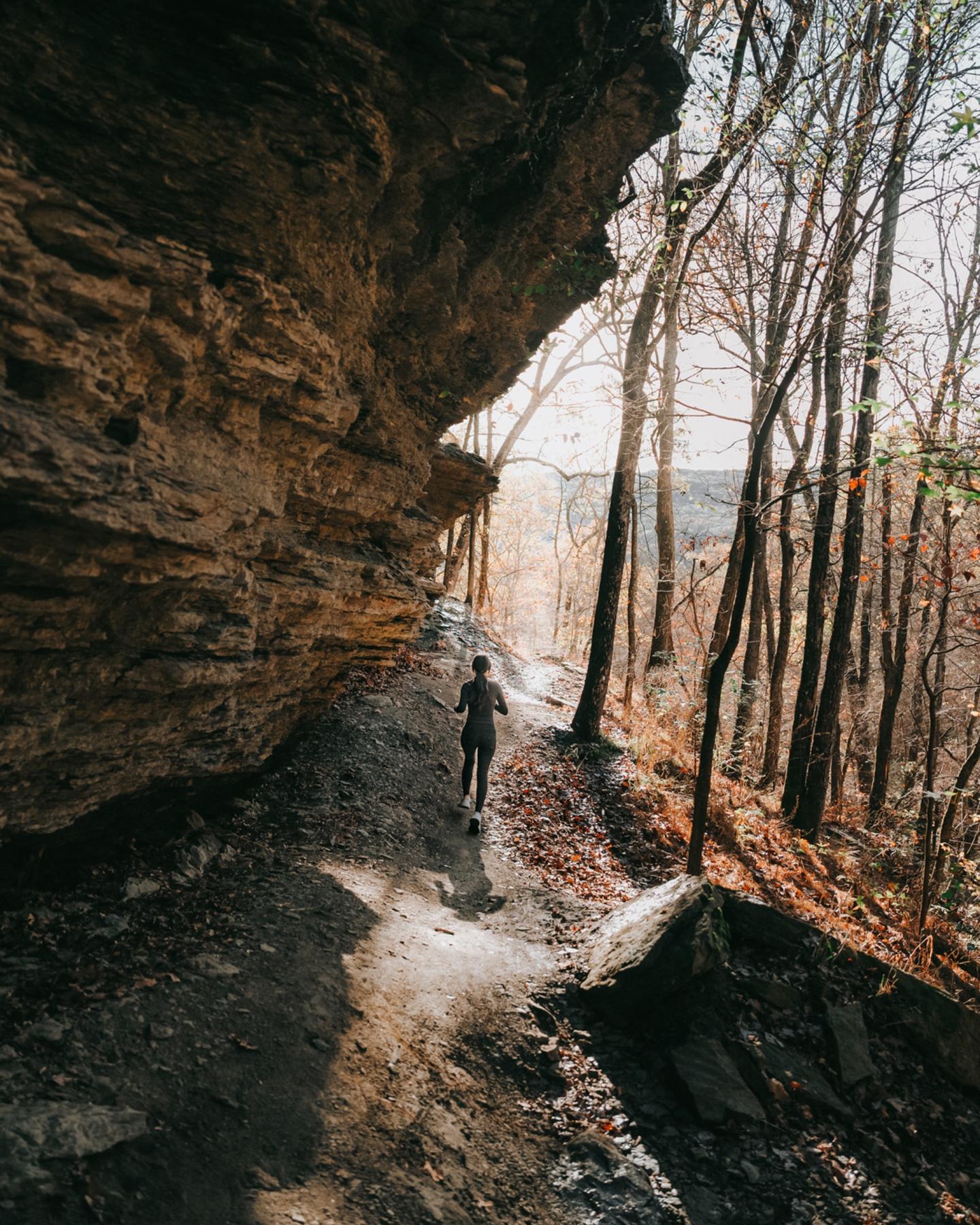 Runner on a forest trail beside a rocky cliff, with winter trees and sunlight filtering through.