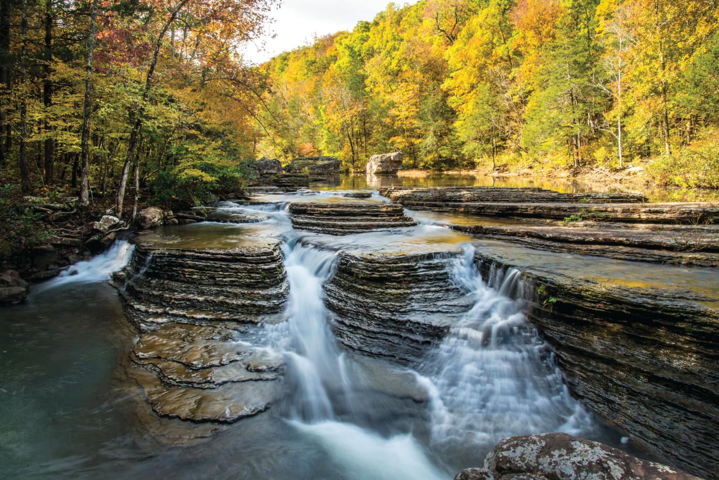 Waterfalls cascading over tiered rock formations, surrounded by autumn trees.