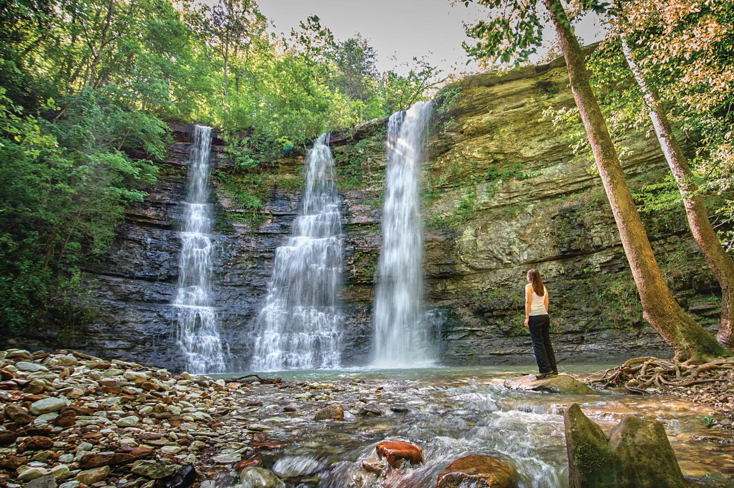 Waterfall with three streams in a lush forest, person standing nearby, serene atmosphere.