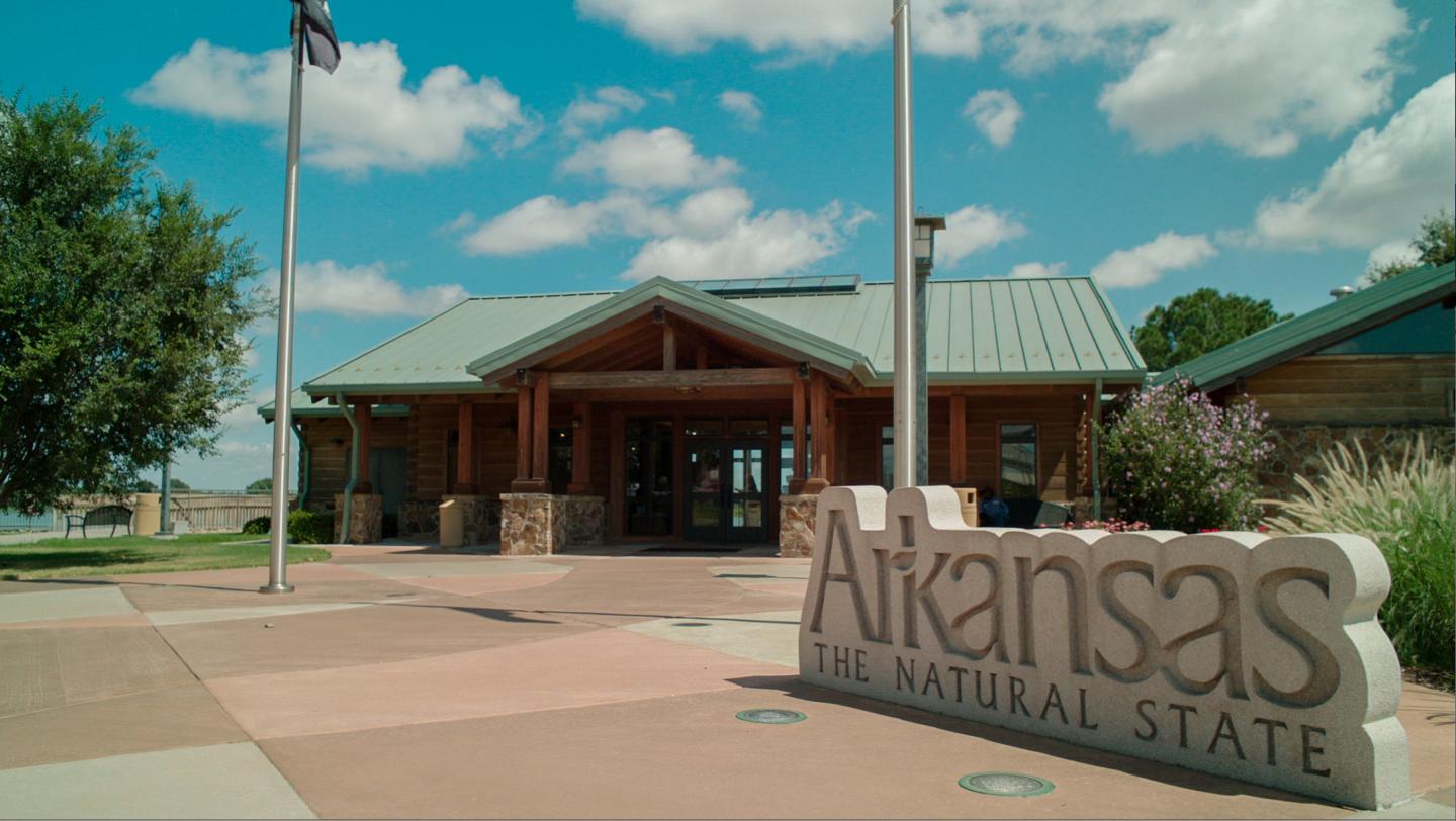 Visitor center with "Arkansas, The Natural State" sign, clear sky, and trees.