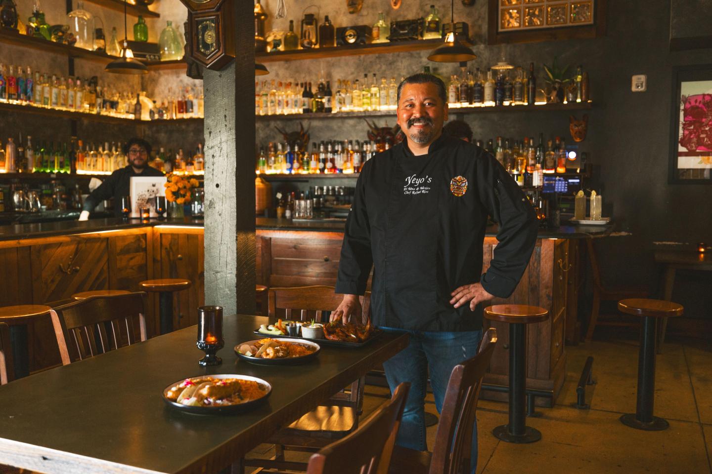 Chef standing in a dimly lit restaurant with plates on a table.