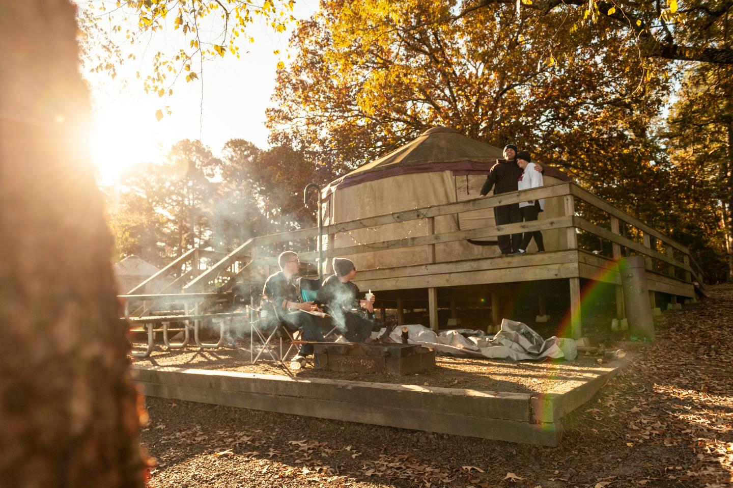 Autumn campsite with people near a fire, wooden cabin, and sunlit trees.
