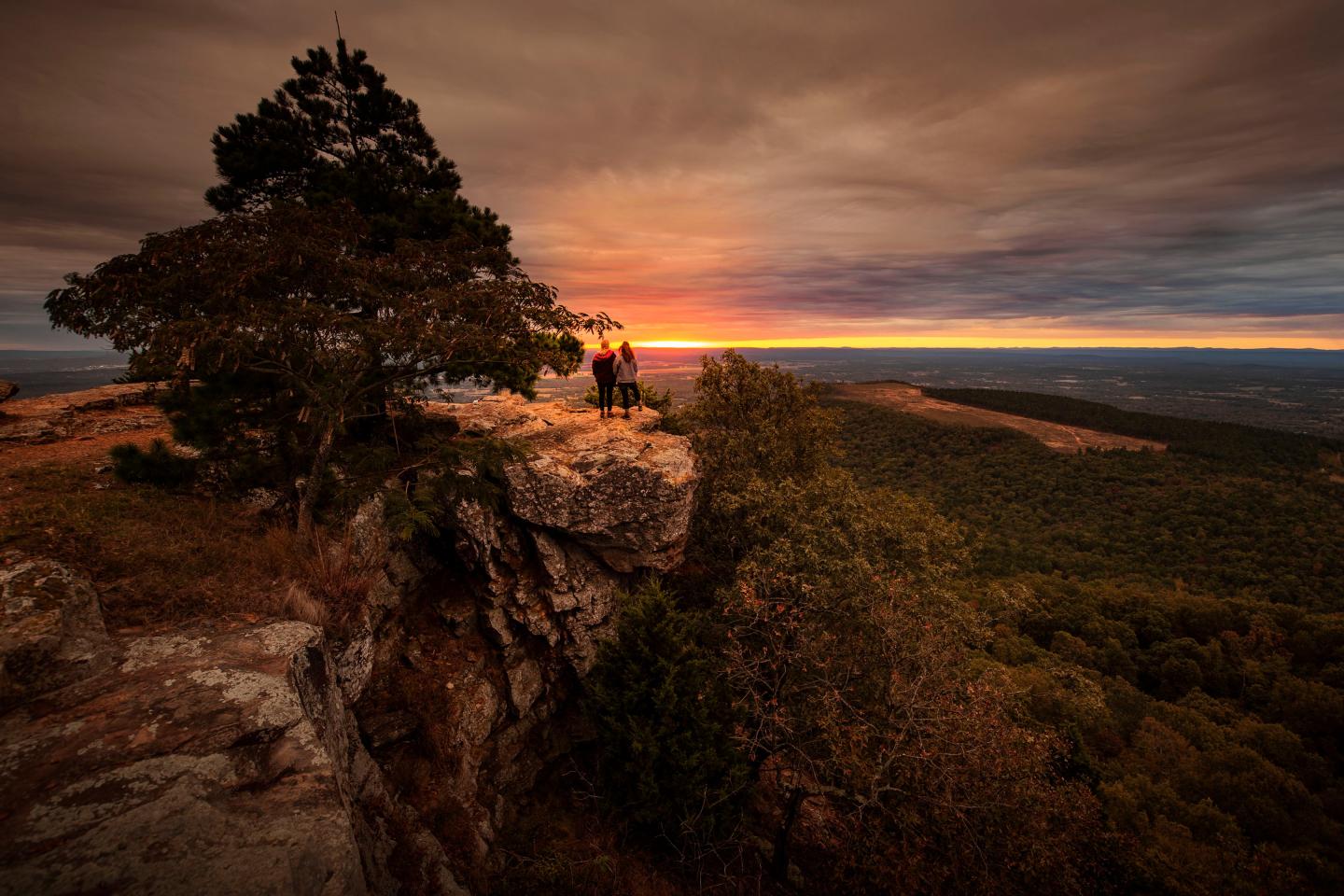 Two people stand on a cliff at sunset, overlooking a vast, green landscape.